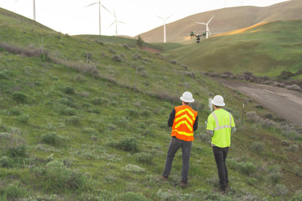 A pair of engineers use a drone to inspect wind farms and power plants in the country. They are standing on a hill and the windmills are a short distance away. There is a rural road at the bottom of the hill.